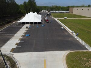 Aerial view of a large white event tent, provided by T3 Event Rentals, set up on an asphalt parking lot in Forsyth County near a grassy area. Several trucks and T3 workers are actively working around the tent. A long beige building is visible to the right with a row of trailers in the background. All items are rentals from T3 Event Rentals, serving Forsyth County and beyond.