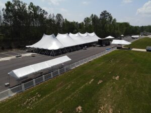 A large white event tent, provided by T3 Event Rentals, stands prominently in a parking lot in Atlanta, flanked by smaller tents and bordered by a white fence. This inviting setup is perfectly placed next to a grassy field with trees under the serene blue sky.