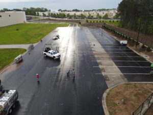 A stunning aerial view showcases a freshly paved parking lot in Dawsonville, featuring freshly painted lines ideal for event rentals. The lot is ready for your next occasion with T3 Event Rentals. Workers and two white trucks can be seen amidst the surrounding trees and nearby industrial buildings. The gleaming pavement indicates recent rainfall or thorough cleaning, enhancing its appeal as an event venue by T3 Event Rentals.
