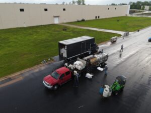 An aerial view of T3 Event Rentals distribution center in Forsyth County showcases a red pickup truck, a green utility vehicle, and a black truck navigating the wet road. Teams are busy loading materials and sacks for event rentals. The backdrop features a large beige building surrounded by lush green grass, highlighting the scenic beauty of Forsyth County.