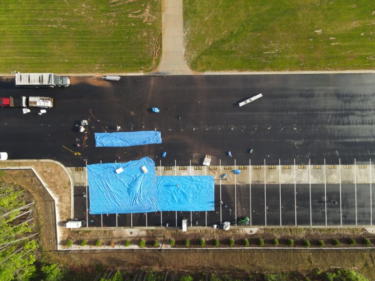 A drone captures a sweeping view of a large parking area in Forsyth County, where two blue tarps are laid out on the asphalt, signaling event preparations by T3 Event Rentals. Surrounding vehicles and rental equipment suggest an upcoming occasion amidst the lush greenery and neatly arranged small shrubs provided by T3 Event Rentals, strategically serving the Forsyth County area for all your event needs.
