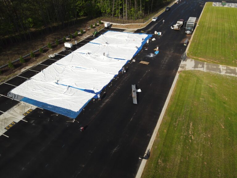 A stunning aerial view captures a massive white and blue tarp laid out in an outdoor parking lot, hinting at the dynamic event rentals in North Georgia. The scene is bustling with people and vehicles surrounding the setup, all courtesy of T3 Event Rentals. This vibrant area is beautifully framed by lush grass and trees, providing a perfect backdrop for any gathering.