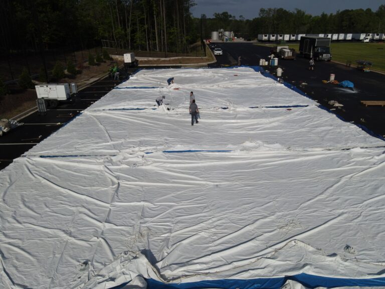 Workers expertly spread a large white tarp across a paved area in Dahlonega, setting the stage for an upcoming event. Encircled by equipment and shipping containers, this scene is framed by trees under a partly cloudy sky—ready to transform this space with top-notch event rentals from T3 Event Rentals. All items on display are available for rent through T3 Event Rentals.
