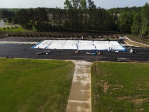 Workers are busily constructing a long, white tent-like structure on a paved area enveloped by grass and trees in Forsyth County, preparing for event rentals. Equipment and rental materials from T3 Event Rentals are scattered around the site, designed to look like a parking lot.