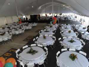 An expansive white tent from T3 Event Rentals graces the scene in Atlanta, sheltering numerous round tables draped with elegant white tablecloths and vibrant tropical centerpieces. Clusters of orange, green, and red balloons are strategically positioned throughout the venue to enhance the festive ambiance. Chairs are neatly arranged around each table, creating an inviting atmosphere for all attendees.