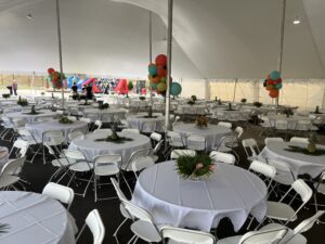 Transform an ordinary gathering into an extraordinary experience with T3 Event Rentals in Forsyth County. Our spacious white tent is equipped with round tables and elegant white folding chairs, setting the stage for a lively event. The tables are enhanced with tropical centerpieces featuring palm leaves and pineapples, while colorful balloons festively decorate the poles. Trust T3 Event Rentals to provide everything needed for a memorable celebration in Forsyth County. Remember, all items are rentals!.