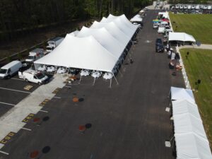Aerial view of a large outdoor event setup with T3 Event Rentals in Forsyth County. Several spacious white tents are arranged in a parking lot, equipped with tables and chairs for attendees. Nearby, food trucks and smaller tents line the sides. The surrounding grassy areas enhance the charm of this vibrant scene.