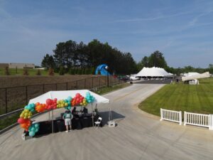 Aerial view of an outdoor event setup in Forsyth County, showcasing vibrant balloon clusters decorating a small canopy with guests beneath. A large white tent and inflatable slide from T3 Event Rentals enhance the scene amidst trees and a lush grassy lawn.