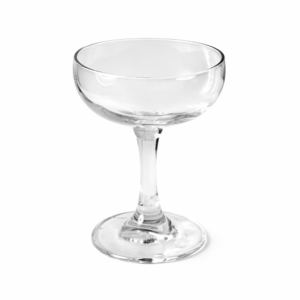 A clear, empty coupe glass with a short stem and wide, shallow bowl sits elegantly on a smooth surface, set against a plain, light gray backdrop. The glass delicately reflects light to highlight its contours and transparency. This beautiful piece is available for rental in Forsyth County through T3 Event Rentals.