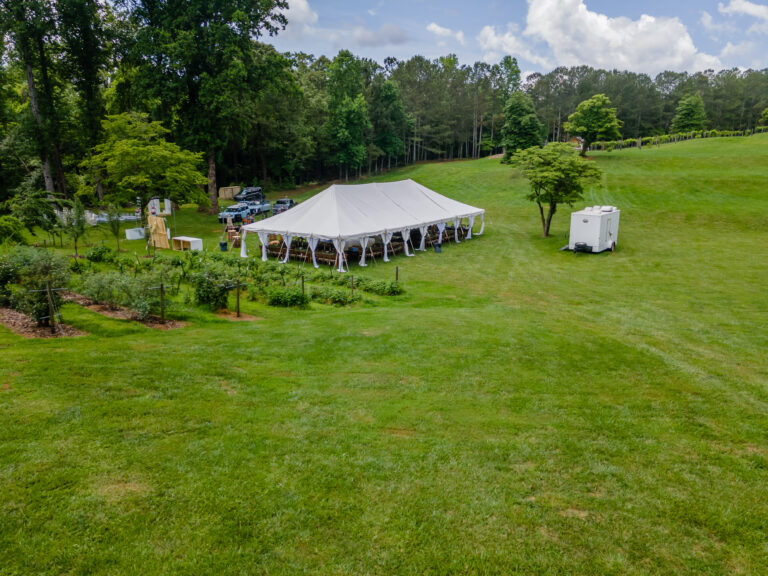 A large white frame event tent from T3 Event Rentals is set up on a grassy field in Ball Ground, surrounded by stylish barrel tables and scattered rental chairs. A small stage—complete with musical instruments—adds a touch of entertainment to the setting. A few guests and parked vehicles can be seen nearby, with trees providing a scenic backdrop. All tents, tables, chairs, and stage equipment are available for rent from T3 Event Rentals in Ball Ground.