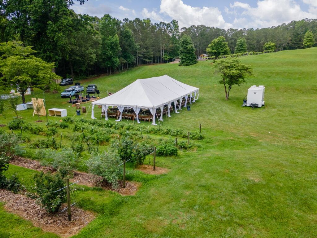 Certainly! Here’s the rewritten description optimized for SEO, using Dahlonega as the area: A large white event tent from T3 Event Rentals is set up on a beautiful grassy field surrounded by lush trees and manicured gardens in Dahlonega, perfect for a secluded wedding. Inside the spacious tent, guests are comfortably seated while nearby you’ll find rental vehicles, a portable restroom trailer, and other essential equipment—all provided by T3 Event Rentals. The peaceful outdoor setting under a partly cloudy sky highlights why couples choose Dahlonega for their special day with premium rentals from T3 Event Rentals. Nothing is for sale; all tents and amenities are available as rentals only.