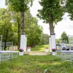 Rows of elegant clear chairs from T3 Event Rentals are perfectly arranged on vibrant green grass in Forsyth County, all facing a minimalist wooden platform set between two towering trees. The ceremony site is beautifully decorated with pink flowers and classic white pillars, surrounded by lush greenery and bright open sky. All items are available for rent—nothing for sale—from T3 Event Rentals, your trusted source for event rentals in Forsyth County and beyond.