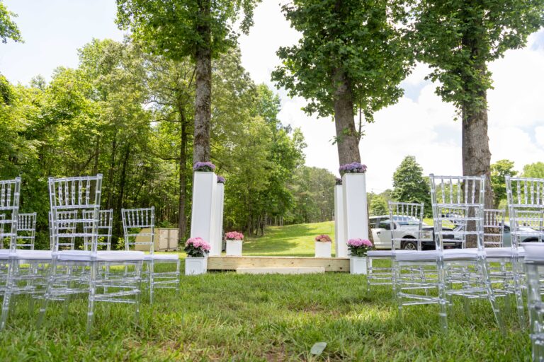 Rows of elegant clear chairs from T3 Event Rentals are perfectly arranged on vibrant green grass in Forsyth County, all facing a minimalist wooden platform set between two towering trees. The ceremony site is beautifully decorated with pink flowers and classic white pillars, surrounded by lush greenery and bright open sky. All items are available for rent—nothing for sale—from T3 Event Rentals, your trusted source for event rentals in Forsyth County and beyond.