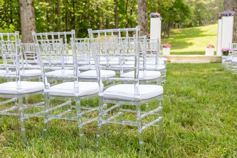 Rows of clear acrylic chairs with white cushions from T3 Event Rentals are set up outdoors on lush green grass, facing a minimalist wedding altar in a beautiful wooded area near Dahlonega. Sunlight peeks through the trees, creating a romantic ceremony space—all rentals provided by T3 Event Rentals for Dahlonega weddings and events.