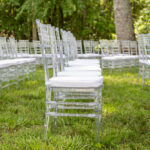 Rows of clear acrylic chiavari chairs with white cushions, perfectly arranged on lush green grass and surrounded by towering trees, create an elegant outdoor event setup in Ball Ground. All items are rentals provided by T3 Event Rentals, serving Ball Ground and the North Georgia area for weddings and special occasions.