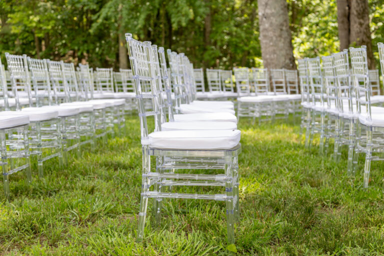 Rows of clear acrylic chiavari chairs with white cushions, perfectly arranged on lush green grass and surrounded by towering trees, create an elegant outdoor event setup in Ball Ground. All items are rentals provided by T3 Event Rentals, serving Ball Ground and the North Georgia area for weddings and special occasions.