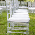 Rows of clear acrylic chairs with white cushions, available for rent from T3 Event Rentals, are neatly arranged outdoors on lush green grass in Forsyth County. Trees and additional seating rentals can be seen in the background, creating an elegant ceremony setup perfect for weddings or special events in Forsyth County. Trust T3 Event Rentals to provide stylish rental seating and event essentials for your next outdoor occasion.