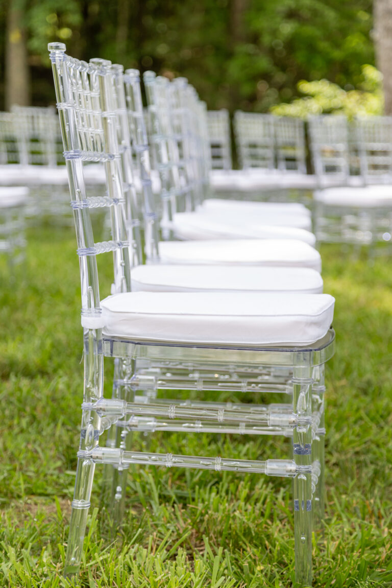 Rows of clear acrylic chairs with white cushions, available for rent from T3 Event Rentals, are neatly arranged outdoors on lush green grass in Forsyth County. Trees and additional seating rentals can be seen in the background, creating an elegant ceremony setup perfect for weddings or special events in Forsyth County. Trust T3 Event Rentals to provide stylish rental seating and event essentials for your next outdoor occasion.