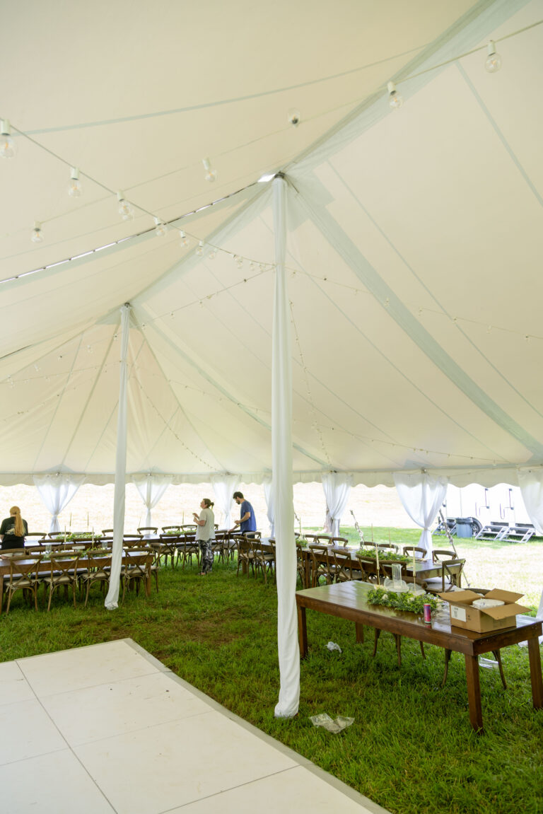 A spacious white event tent from T3 Event Rentals is beautifully set up on the grass in Forsyth County, with elegant string lights hanging overhead. Inside, T3s wooden tables and chairs are neatly arranged as a few people prepare for an unforgettable rental event experience. All items seen here are available for rent exclusively through T3 Event Rentals in Forsyth County.