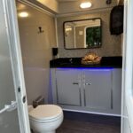 For events in Atlanta, T3 Event Rentals provides the 2 Stall Restroom Trailer—a clean and modern rental featuring white toilets, gray cabinets, sleek black countertops, wall mirrors, rolled towel baskets, and ambient blue lighting under the counter for a bright and inviting atmosphere. Trust T3 Event Rentals to elevate your next Atlanta gathering with this stylish restroom trailer. Let me know which location youd like a version for next!.