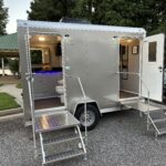 A 2 Stall Restroom Trailer from T3 Event Rentals is shown onsite in Dawsonville, featuring two open doors with sturdy metal steps and railings. This rental unit is positioned conveniently on pavement adjacent to grass and trees. Inside the restroom trailer, amenities include sinks, a spacious countertop, and visible toilets—perfect for your next event in Dawsonville. T3 Event Rentals proudly serves the Dawsonville area with high-quality restroom trailer rentals.