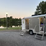 T3 Event Rentals offers a 2 Stall Restroom Trailer for rent in Dahlonega, perfect for outdoor gatherings and special events. This portable restroom trailer is shown parked by a scenic lake at sunset, both doors open to reveal clean interiors. Sturdy metal steps provide safe access at each entrance. The setting features trees, a lamppost in the background, and a rental bench right near the water’s edge—ideal for weddings or events in Dahlonega. Trust T3 Event Rentals to provide high-quality restroom trailers and event rentals throughout North Georgia.